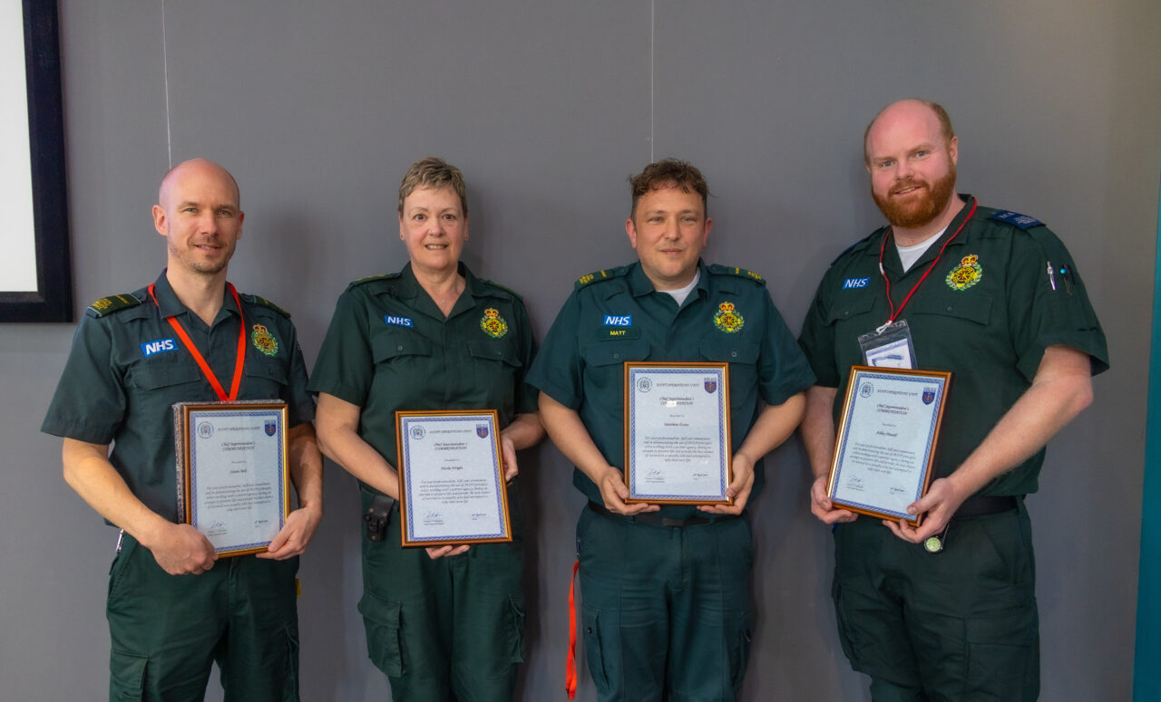 Four ambulance staff in green uniforms standing side by side holding a framed commendation each