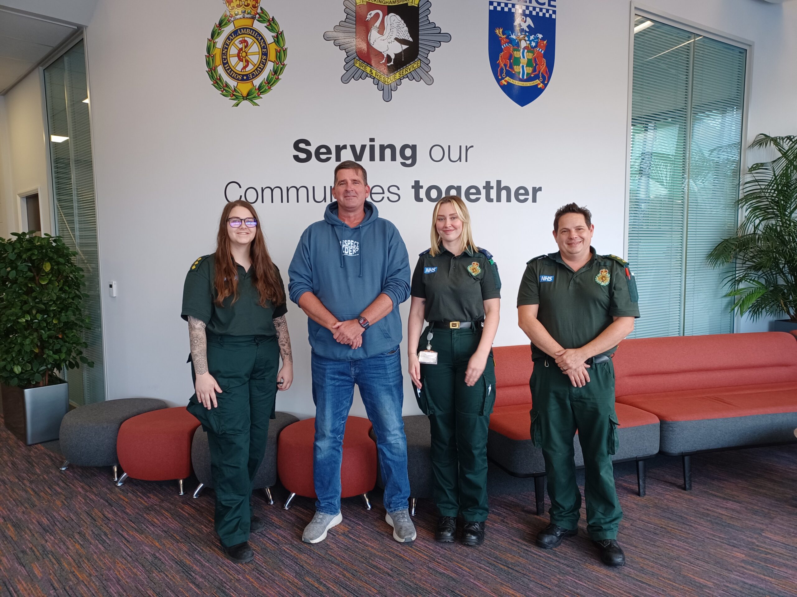 Three ambulance staff in green uniforms standing with a member of the public in front of a sign that reads Serving our Communities together in the reception area of the Blue Light Hub in Milton Keynes