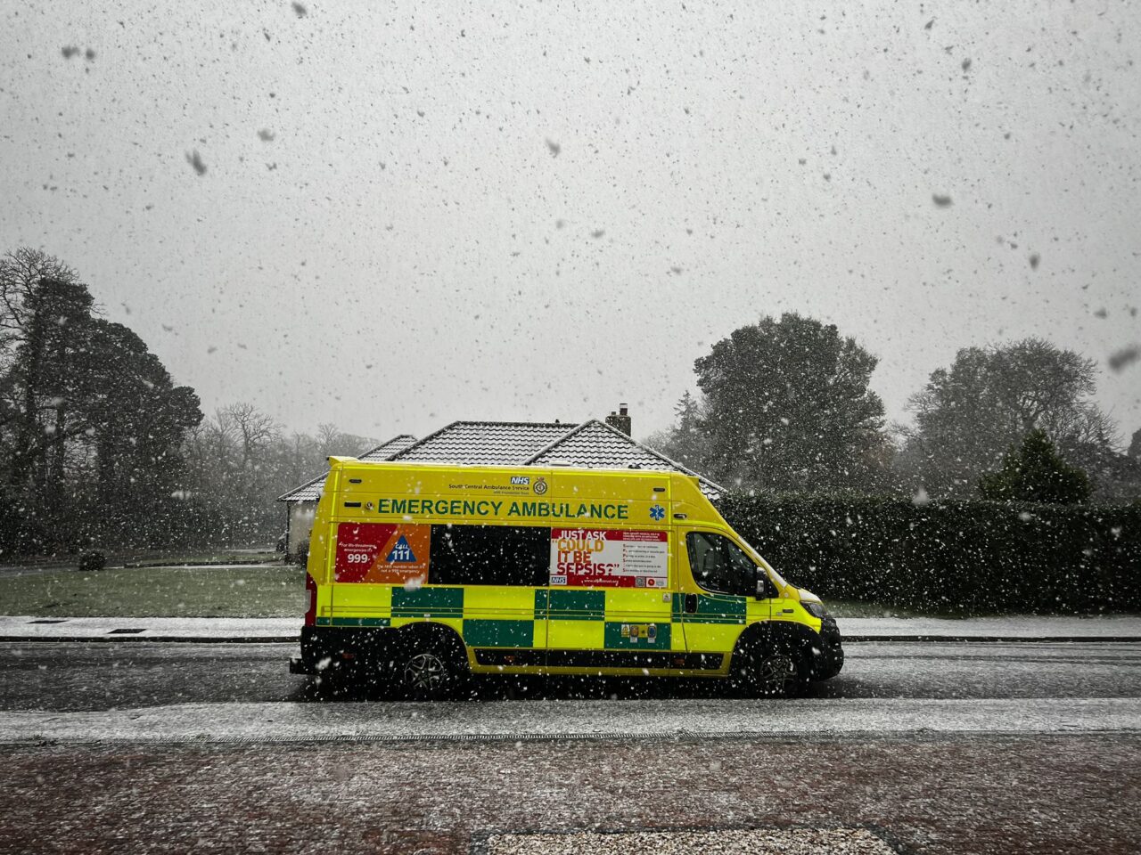 An ambulance parked in front of a house whilst it is snowing.