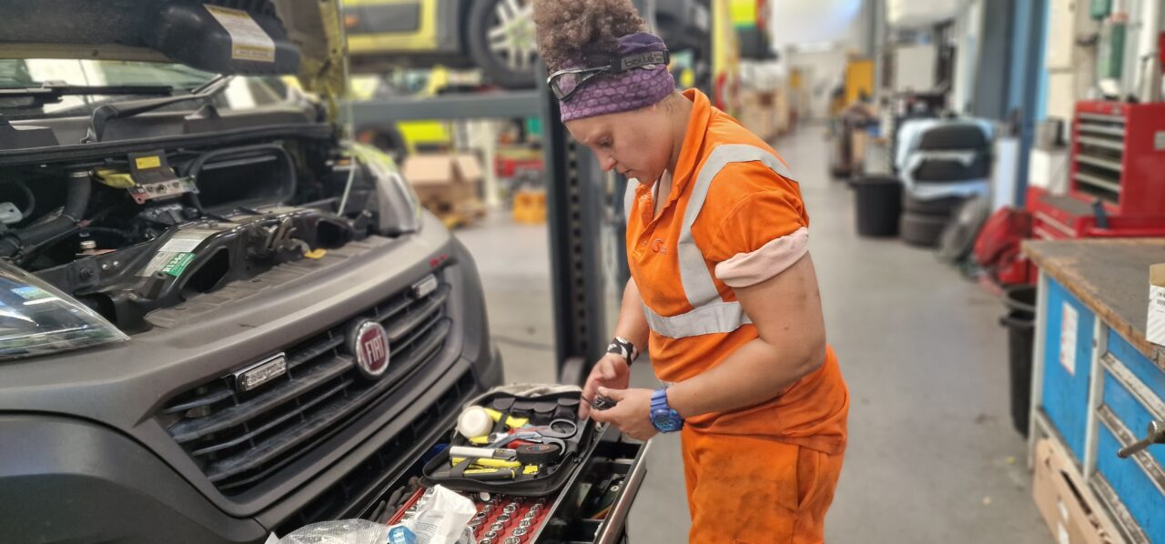 A vehicle technician in a garage working on an ambulance which has its front hood up exposing the engine